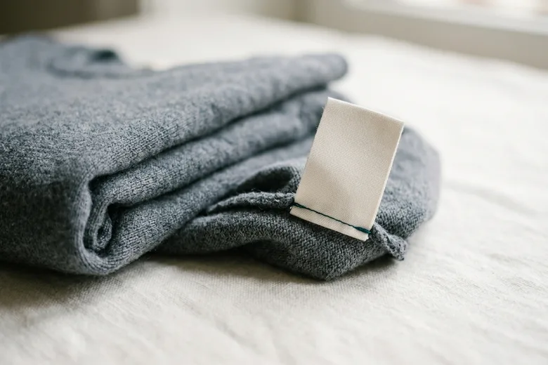 Ein eingenähtes Pflegeetikett ragt aus der Naht eines gefalteten Strickteils; die bedruckte Seite ist vollständig abgewandt.
